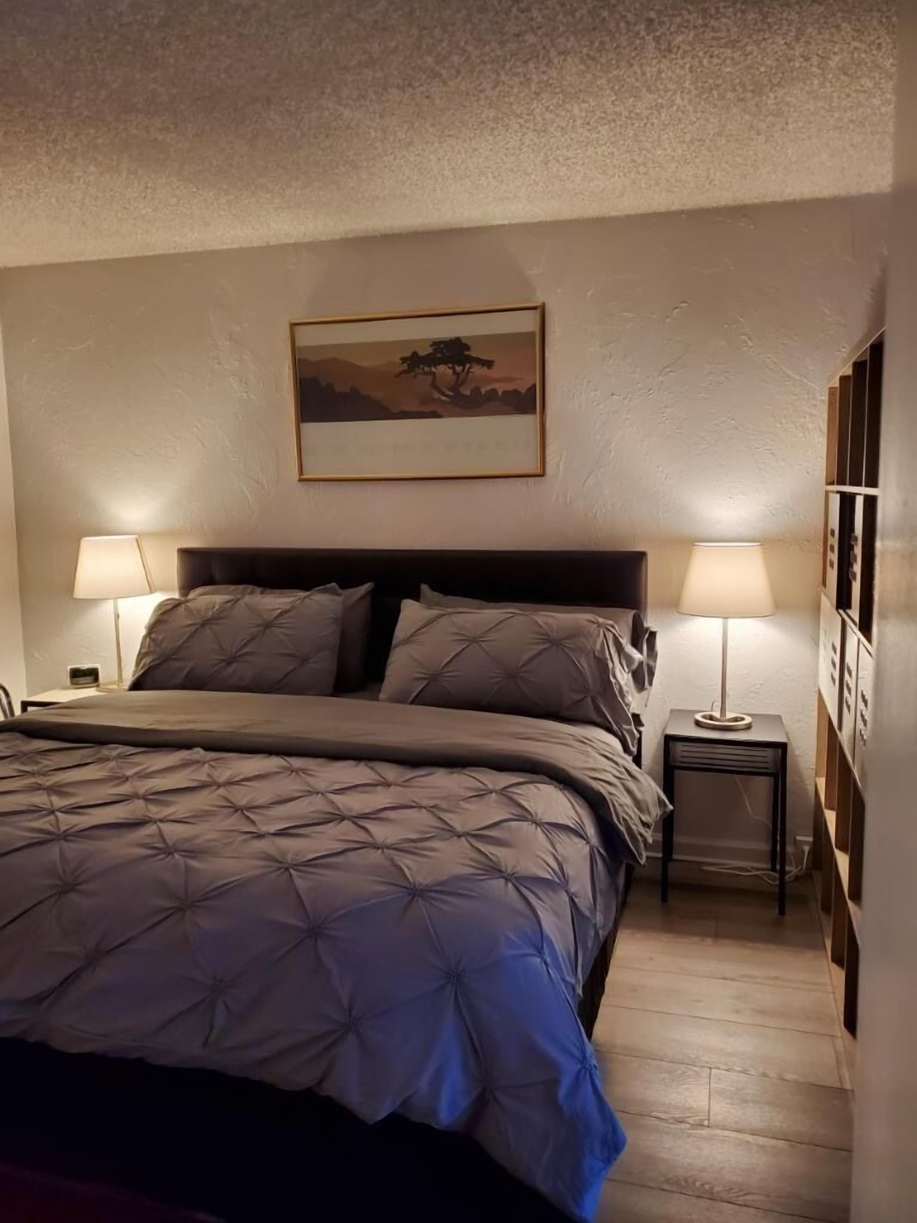 A bedroom with a bed, two bedside tables with lamps, a wall-mounted picture, and a wooden floor.