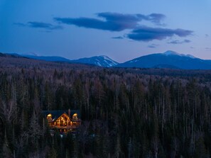 A lodge with a large deck and multiple windows, surrounded by a forest and mountains in the background. - River Road Loj - Lake Placid log cabin rental (Lake Placid)