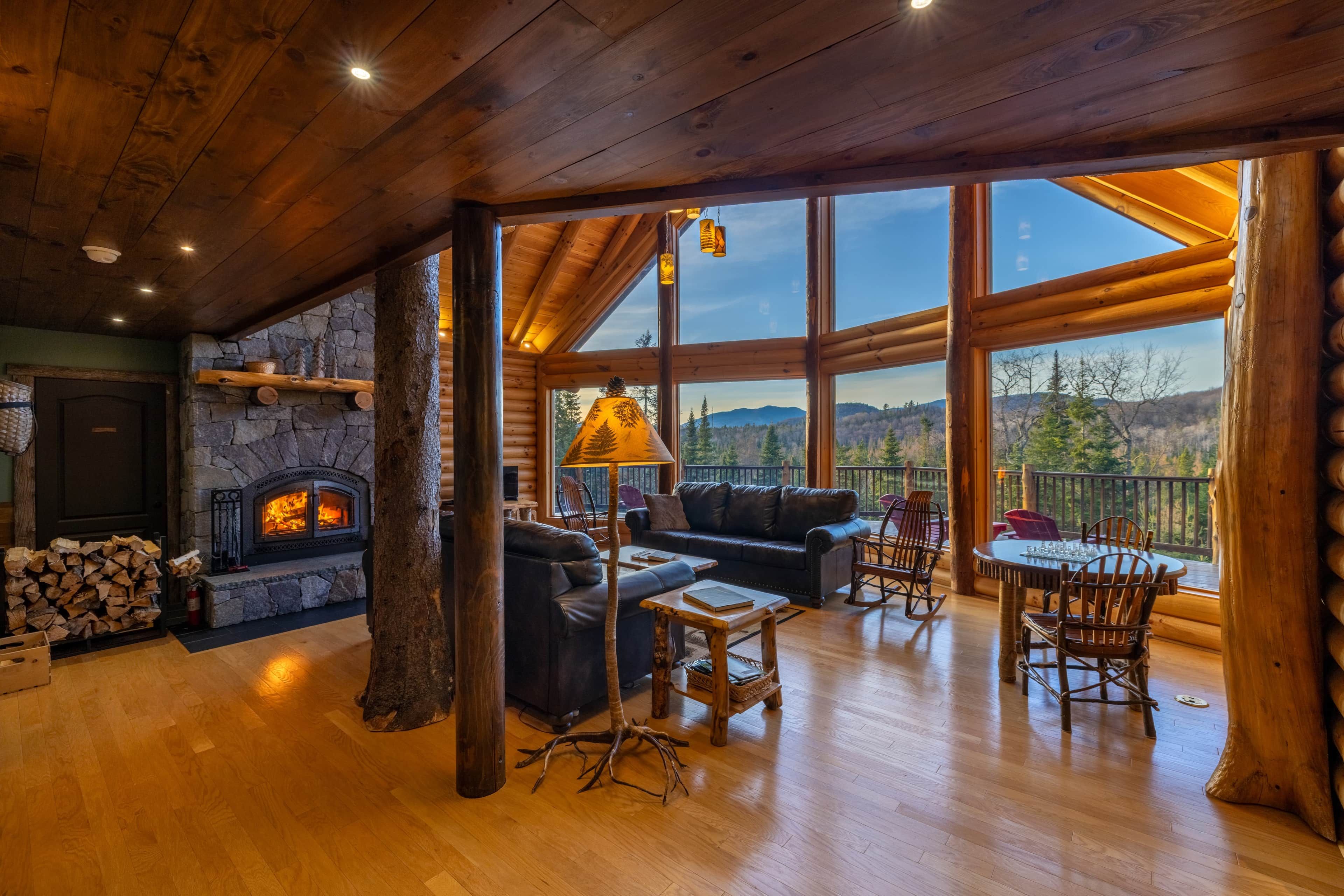 A spacious log cabin interior with a stone fireplace, leather seating, and a balcony with a scenic mountain view.