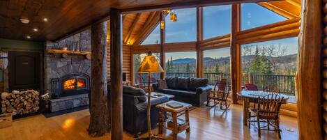 A spacious log cabin interior with a stone fireplace, leather seating, and a balcony with a scenic mountain view.