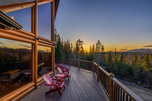 A wooden deck with red Adirondack chairs and a glass enclosure overlooking a forested landscape during sunset. - River Road Loj - Lake Placid log cabin rental (Lake Placid)
