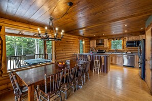 A log cabin interior with a dining area, kitchen, and a view of the forest. - River Road Loj - Lake Placid log cabin rental (Lake Placid)
