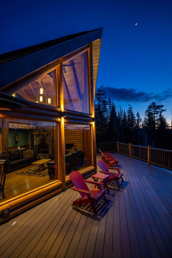 Une terrasse en bois avec des fauteuils Adirondack rouges et une pièce fermée par des baies vitrées, dotée d’une cheminée.