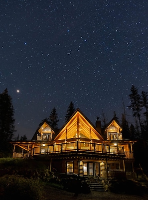 Une cabane en bois dotée d’un balcon, entourée d’arbres, sous un ciel étoilé.