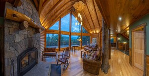Het interieur van een blokhut met een stenen openhaard, houten plafond en grote ramen met uitzicht op het omliggende landschap.