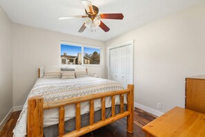 Ein Schlafzimmer mit einem hölzernen Bett, einem Deckenventilator, einem Fenster mit Blick auf Häuser und einer weißen Tür.