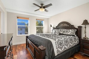 Une chambre avec un grand lit, une tête de lit en bois, deux fenêtres équipées de stores, un ventilateur de plafond et une table de chevet avec une lampe.
