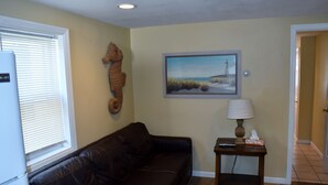 A living room with a dark leather couch, a small table with a lamp, a framed painting of a coastal scene, and a wooden seahorse decoration.
