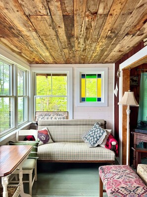 A cozy room with a wooden ceiling, a bench with cushions, a table, and a window with a view of trees.