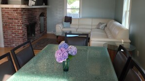 Una sala de estar con chimenea de ladrillo, un sofá blanco, una mesa de centro de vidrio y una mesa de comedor con sillas.