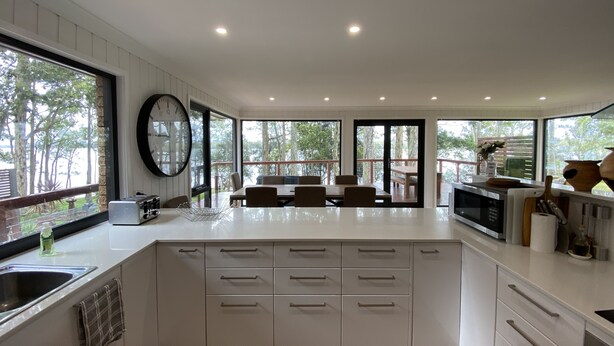 A modern kitchen with white cabinets, a large island, and a dining area with a view of the outdoors. - Premium Waterfront - Jervis Bay Area (St Georges Basin)