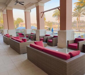 Un espace extérieur aménagé, abrité et doté de mobilier en osier et de coussins rouges, offrant une vue sur une piscine et des palmiers.