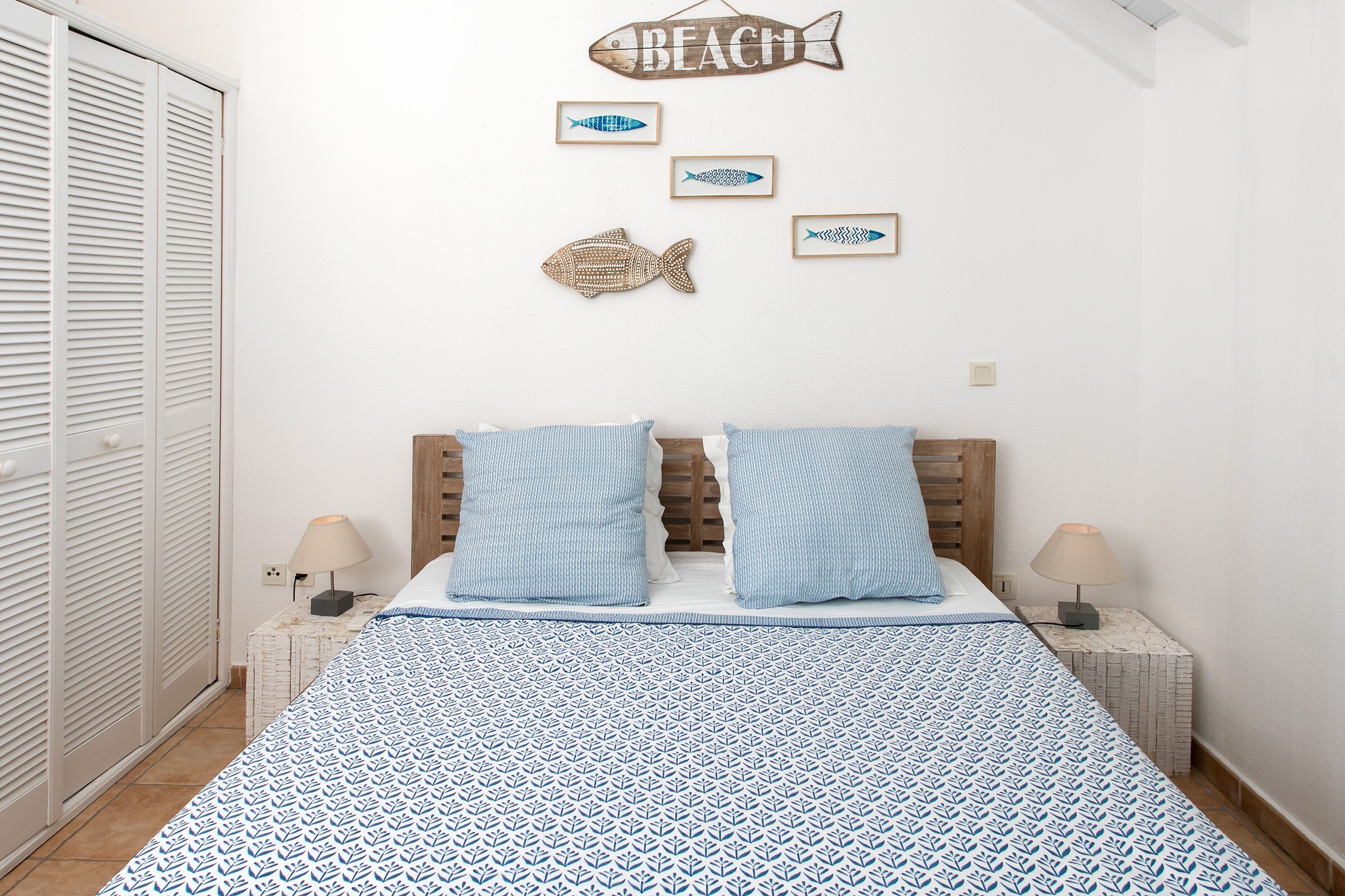 A bedroom with a bed, two bedside lamps, a wooden headboard, a wall with framed fish art, and a closet.