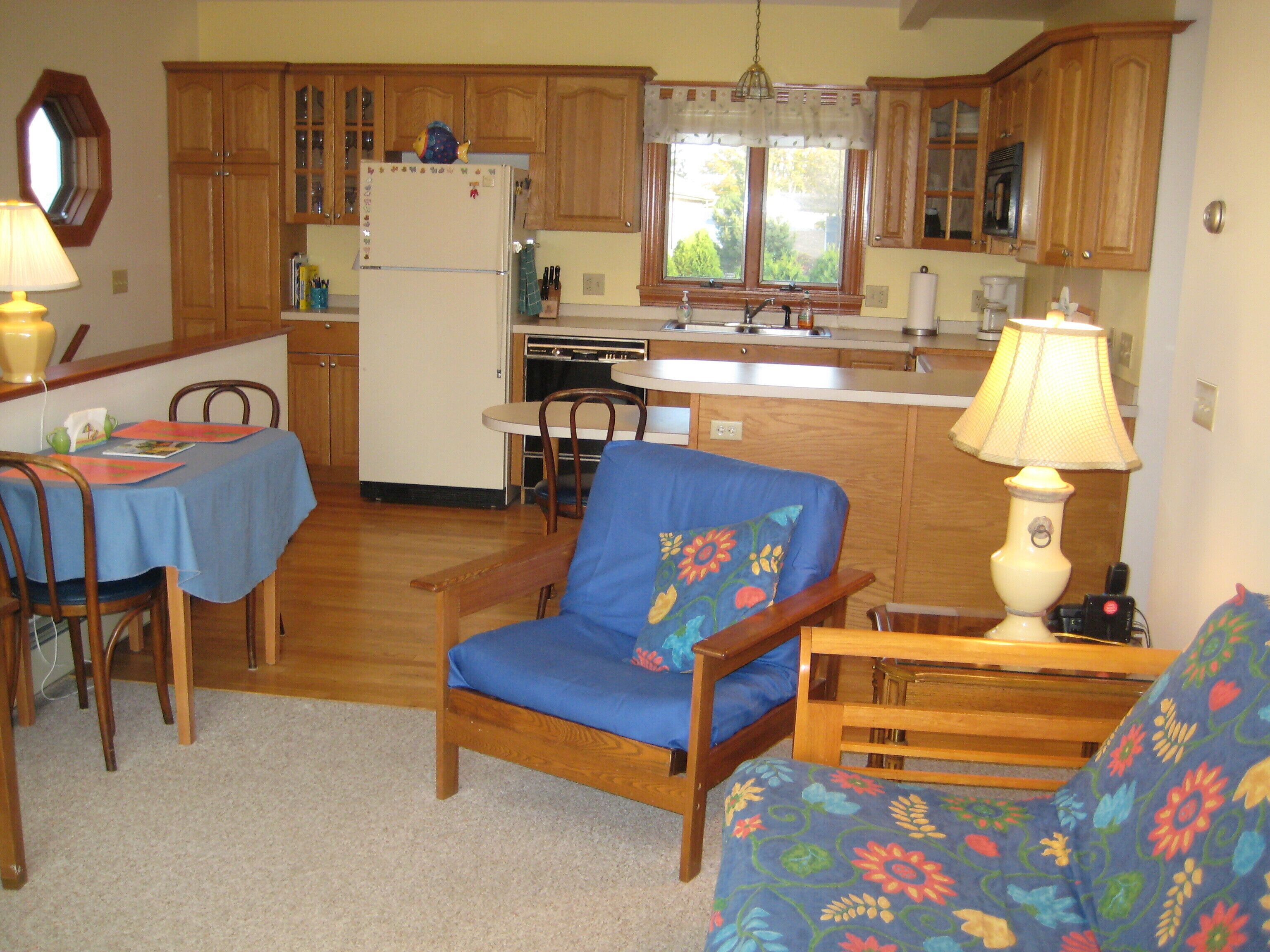 Una cocina compacta con armarios de madera, un refrigerador, una mesa de comedor con sillas y un sillón azul.