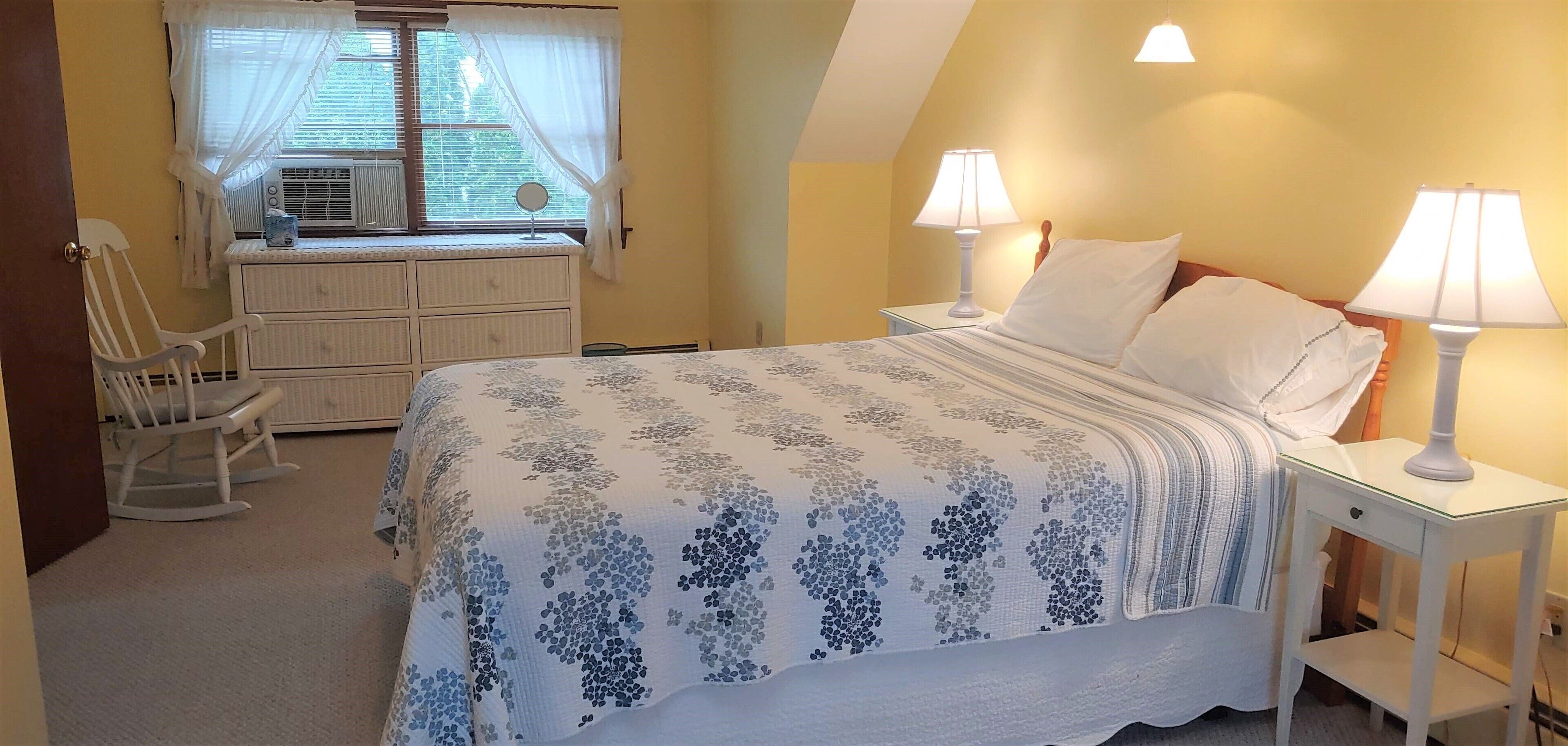 Un dormitorio con cama, dos mesitas de noche con lámparas, cómoda, mecedora y ventana con cortinas.