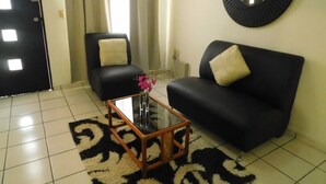 Un salon avec un canapé noir, un fauteuil noir, une table basse en verre ornée d’un vase de fleurs et un grand miroir rond noir accroché au mur.