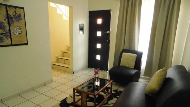 Un salon avec un canapé noir, une table basse en verre et un tapis à motifs. On aperçoit une porte avec un interrupteur et un escalier au fond.