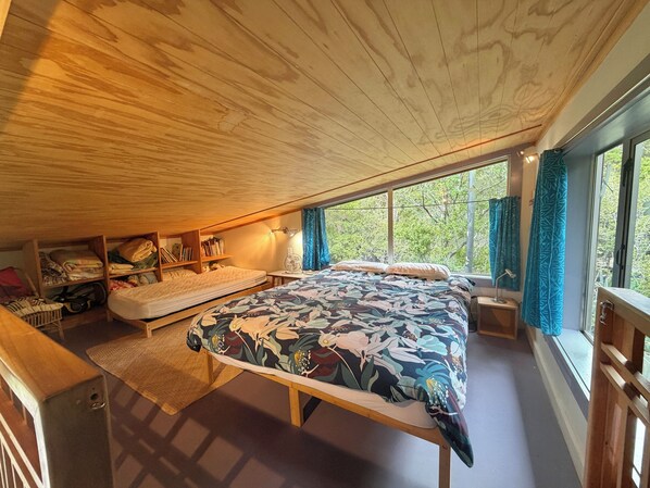 A bedroom with a large bed, wooden ceiling, built-in shelves, a bedside table with a lamp, and a view of trees through large windows. - The Library Bach - A special place people love! (Pakawau Beach)