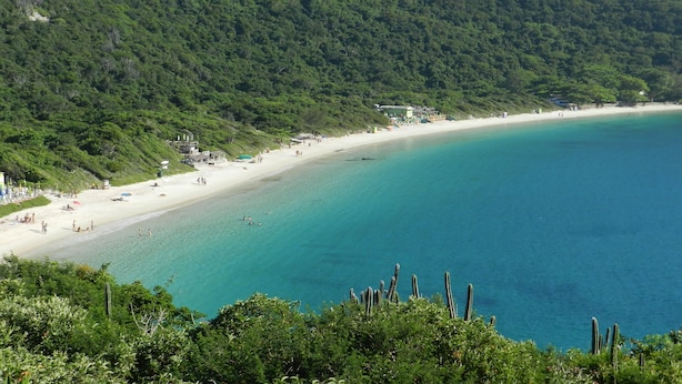 Ein Küstenstrand mit sandigem Ufer, klarem türkisblauem Wasser und grünen Hügeln im Hintergrund.