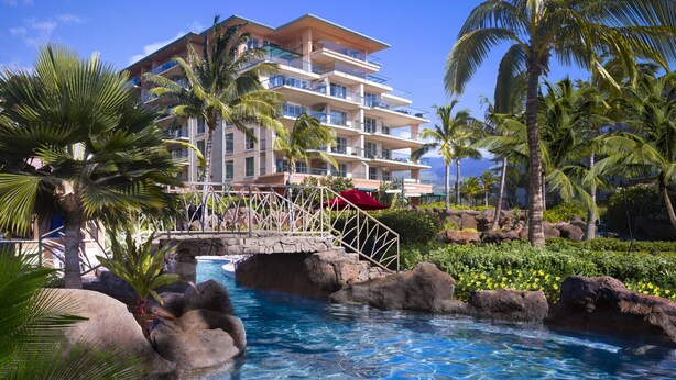 Ein Resort mit Pool, umgeben von Felsen und Palmen, ein mehrgeschossiges Gebäude mit Balkonen und ein strahlend blauer Himmel.