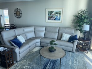 A living room with a large L-shaped sectional sofa, a small round coffee table with a plant, a framed painting on the wall, and a potted plant by the window.
