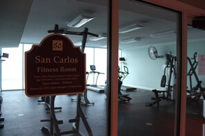 A fitness room with exercise equipment and a sign detailing the room's name, attire requirements, age restriction, and operating hours.