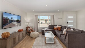 A modern living room with a gray sofa, a coffee table, a TV mounted on the wall, and a large window with a view of a house. - Quiet Scottsdale Retreat ~ 3BR ~ FirePit, Garage, EV Charging ~ Prime Location (Scottsdale)