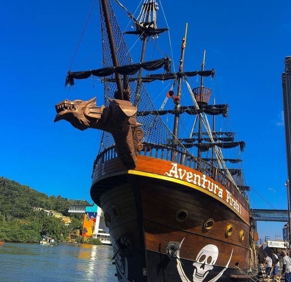 Un barco pirata llamado "Aventura Pirata" atracado en un puerto con cielo azul despejado y un puente al fondo.