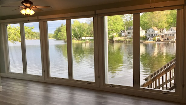 A room with large windows overlooking a lake, a ceiling fan, and a wooden deck.