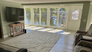 A living room with a large flat-screen TV, a wooden entertainment unit, a patterned rug, and a view of a water body through large glass doors.