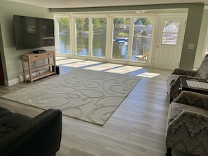 A living room with a large flat-screen TV, a wooden entertainment unit, a patterned rug, and a view of a water body through large glass doors. - ~Baboosic Lake Waterfront Rental~ (Amherst)