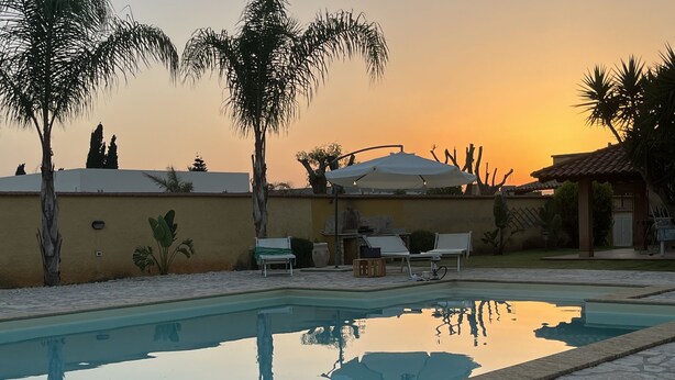 Una piscina con lettini e ombrelloni, circondata da palme e con il sole al tramonto.