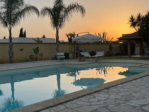 Une piscine entourée de chaises longues et de parasols, bordée de palmiers et offrant une vue sur le coucher du soleil.