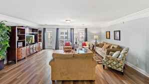 Un salón amplio con un sofá grande, una mesa de centro y un sillón estampado. Se observa un mueble de madera con puertas de cristal y una planta en maceta.