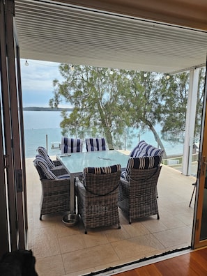 A glass-enclosed patio with wicker furniture and a table, overlooking a body of water and trees.