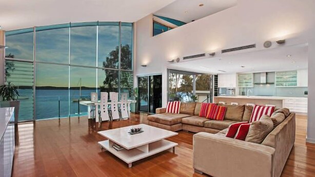 Amplia sala de estar con un gran ventanal con vistas a un cuerpo de agua, cocina moderna y una zona de estar con una mesa de centro blanca y sofás.