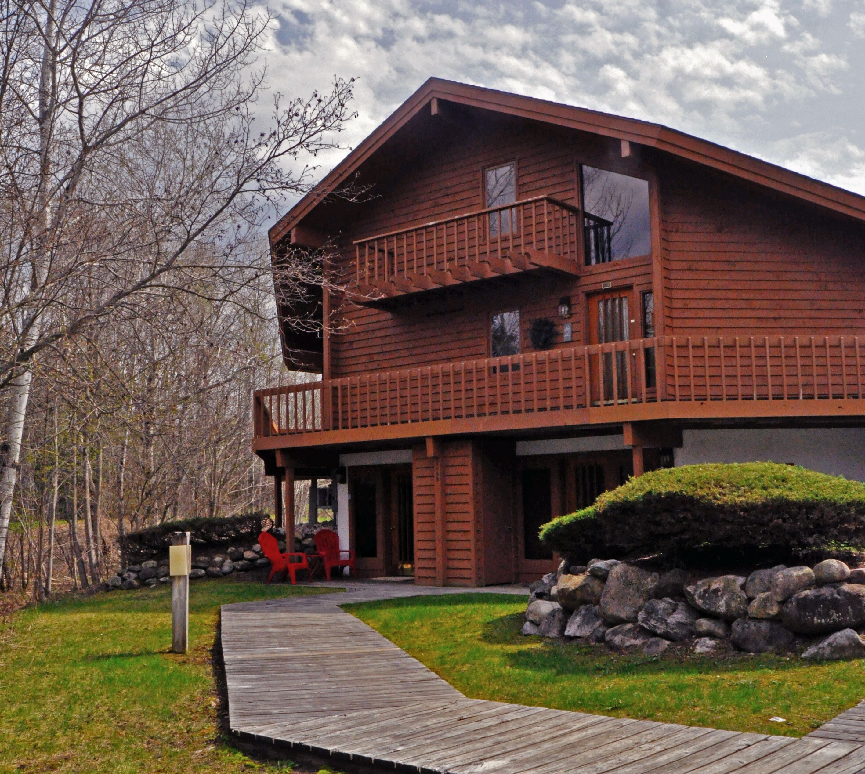 A two-story wooden house with a balcony, a wooden deck, and a stone wall.