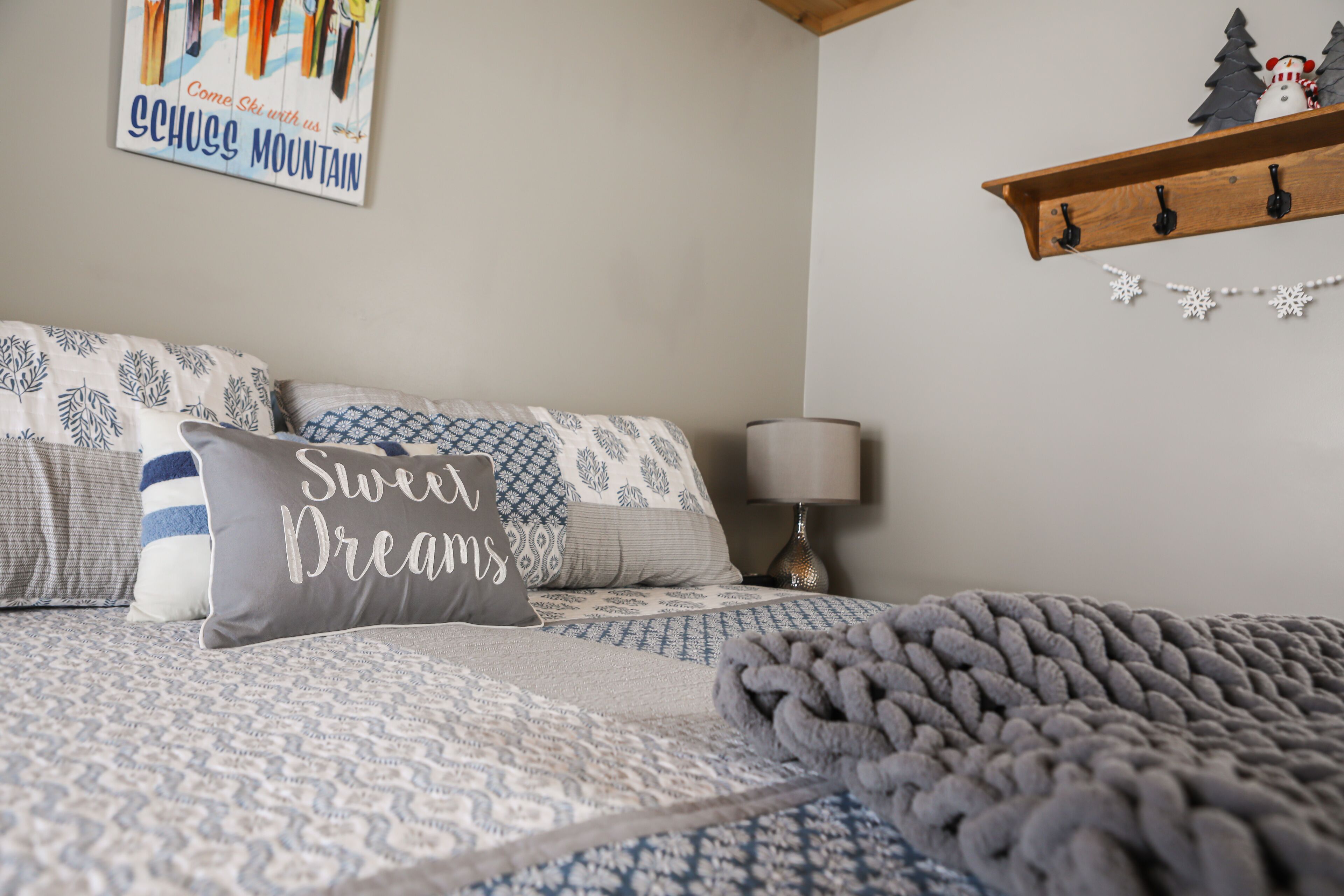 A bedroom with a patterned bedspread, multiple pillows, a grey blanket, a wall-mounted shelf with a snowman decoration, and a sign above the bed.