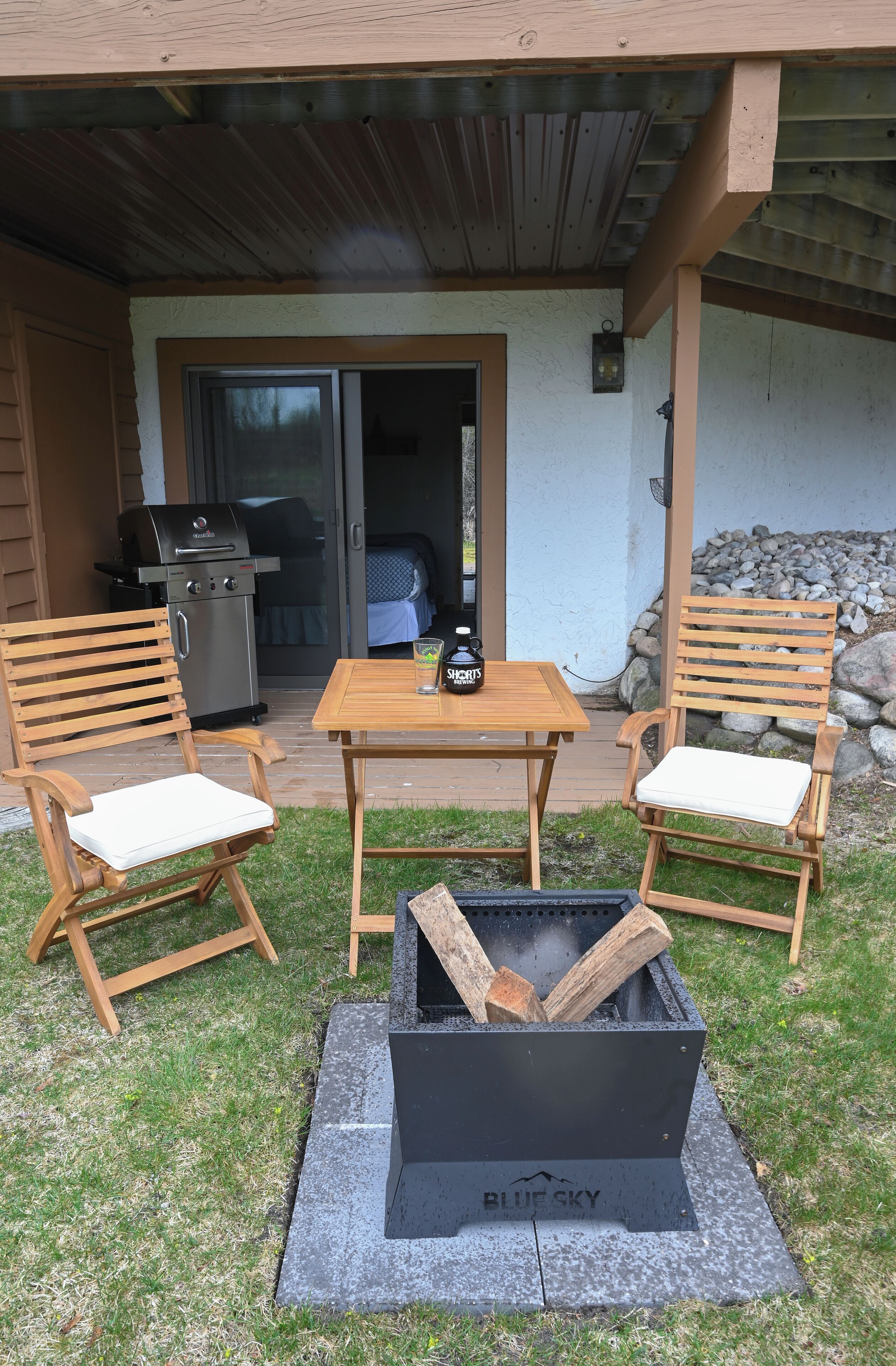 Een terras buiten met twee houten stoelen, een tafel en een zwarte vuurkorf met de tekst 'BLUE SKY'.