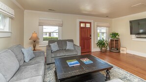 A living room with a grey sofa, a coffee table with books, a wooden floor, a TV, and a door with a window.