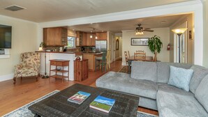 A spacious living room with a grey sofa, a coffee table with books, wooden flooring, a kitchen area with wooden cabinets, and a dining area with a table and chairs.