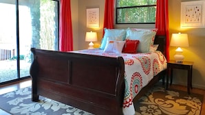 A bedroom with a large bed, two bedside lamps, a ceiling fan, and a window with red curtains.