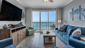 Ein geräumiges Wohnzimmer mit einem großen Flachbildfernseher, einem Deckenventilator, einem Holztisch, einem blauen Sofa und Blick auf den Ozean durch große Fenster.