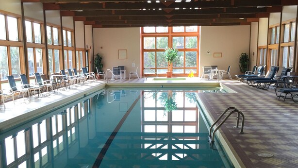 Une piscine intérieure à l’eau claire, entourée de chaises longues et de transats, dotée de grandes fenêtres et d’une structure de plafond en bois.