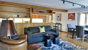 A modern living room with a sofa, coffee table, and chairs. There is a dining area with a table and chairs. The room has wooden beams on the ceiling and a large window. - Immaculate Updated Condo, 5 Minutes from Skiing!  (Wilson)