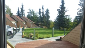 Blick von einem Balkon auf einen gepflegten Garten mit einer Holzterrasse, einer Rasenfläche und einer Reihe von Häusern mit geneigten Dächern.
