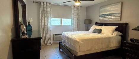 A bedroom with a bed, a chair, a ceiling fan, a window with curtains, and a painting on the wall.