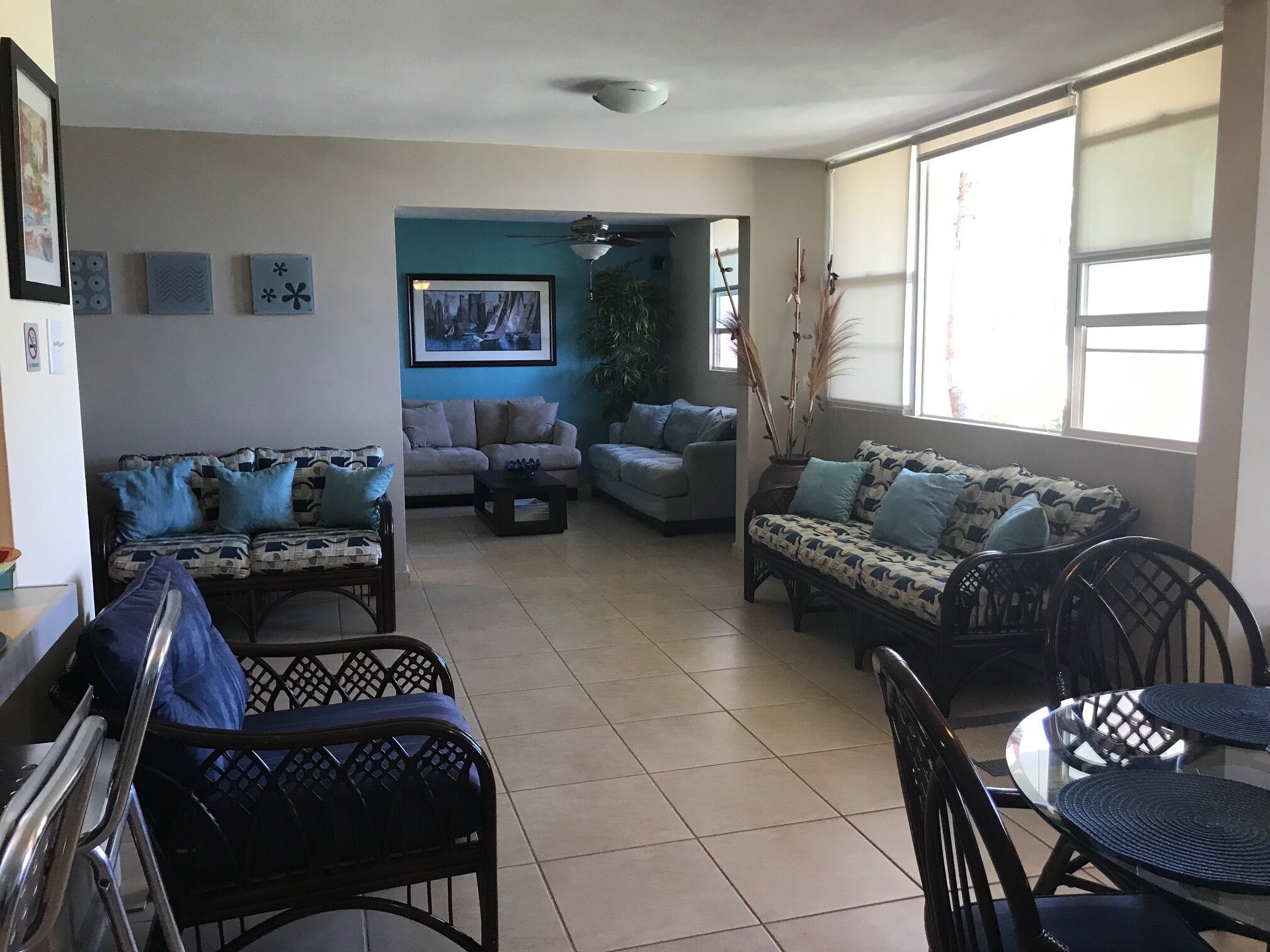 Un salon comprenant deux canapés, une table basse et une table à manger avec des chaises. Les murs sont décorés et il y a un ventilateur de plafond.