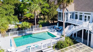 A pool with a wooden deck, surrounded by white railings and blue chairs, with a building in the background. - Sand Castle | Heated Saltwater Pool, Game Room & Walk to Beach and Center Street (Folly Beach)
