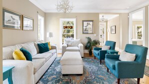 A living room with a beige sofa, blue armchairs, a white ottoman, and a patterned rug. - Sand Castle | Heated Saltwater Pool, Game Room & Walk to Beach and Center Street (Folly Beach)